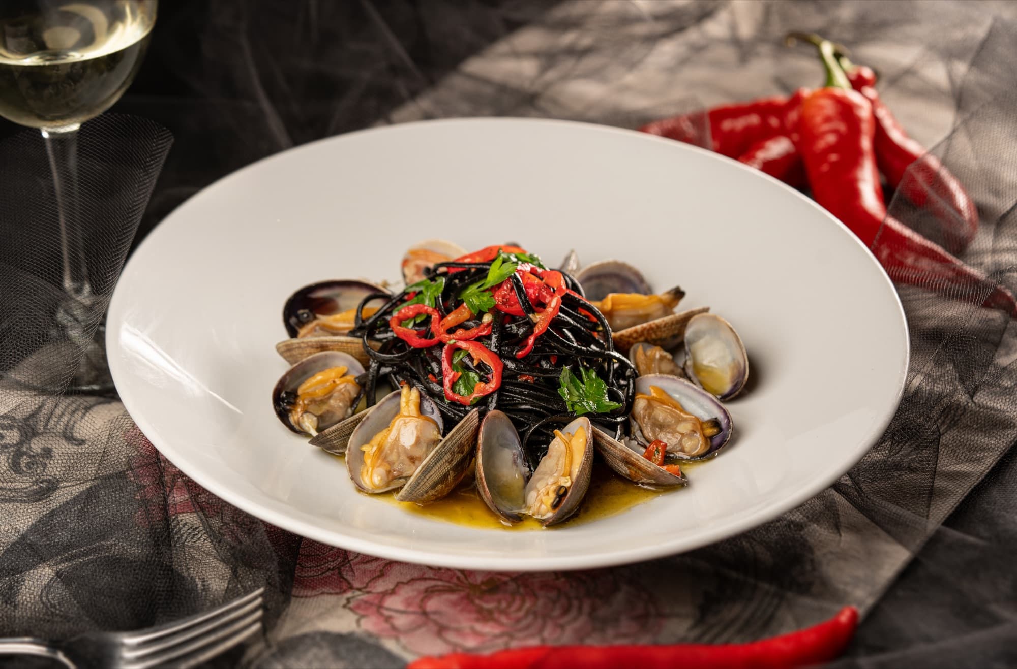 Squid ink spaghetti with clams, red chilli and parsley in a white bowl, beside white wine and fresh chillies.