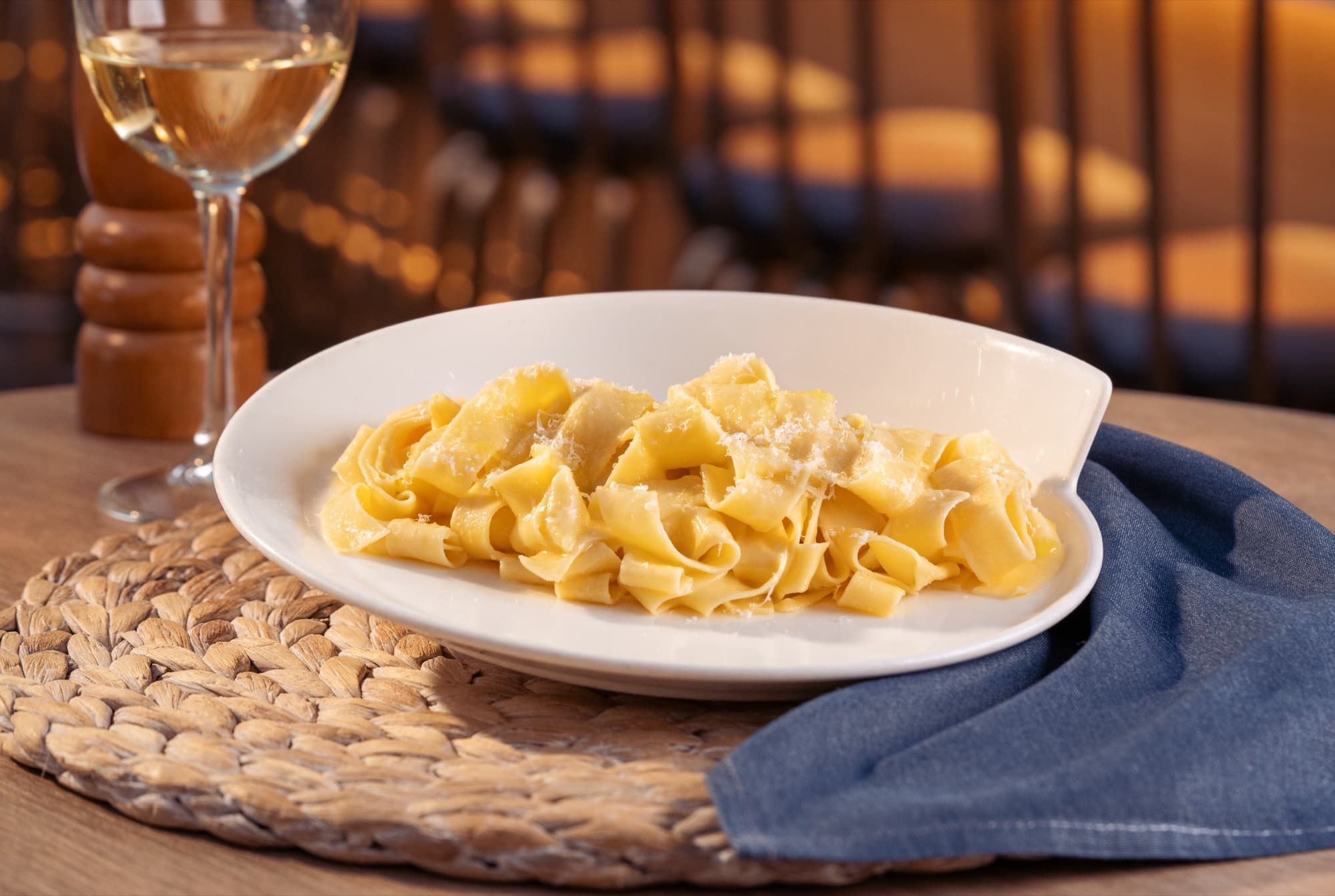 Wide ribbons of pappardelle dusted with grated cheese on a white oval plate beside a glass of white wine.